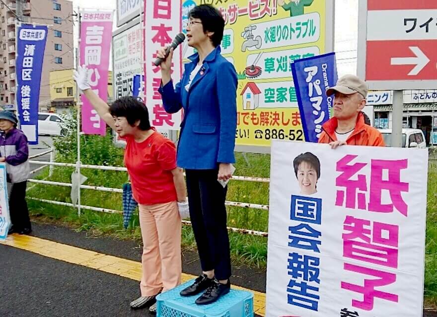 n>「政治を前向きに変える力は日本共産党」と訴える紙議員と聴衆に手を振る吉岡さん